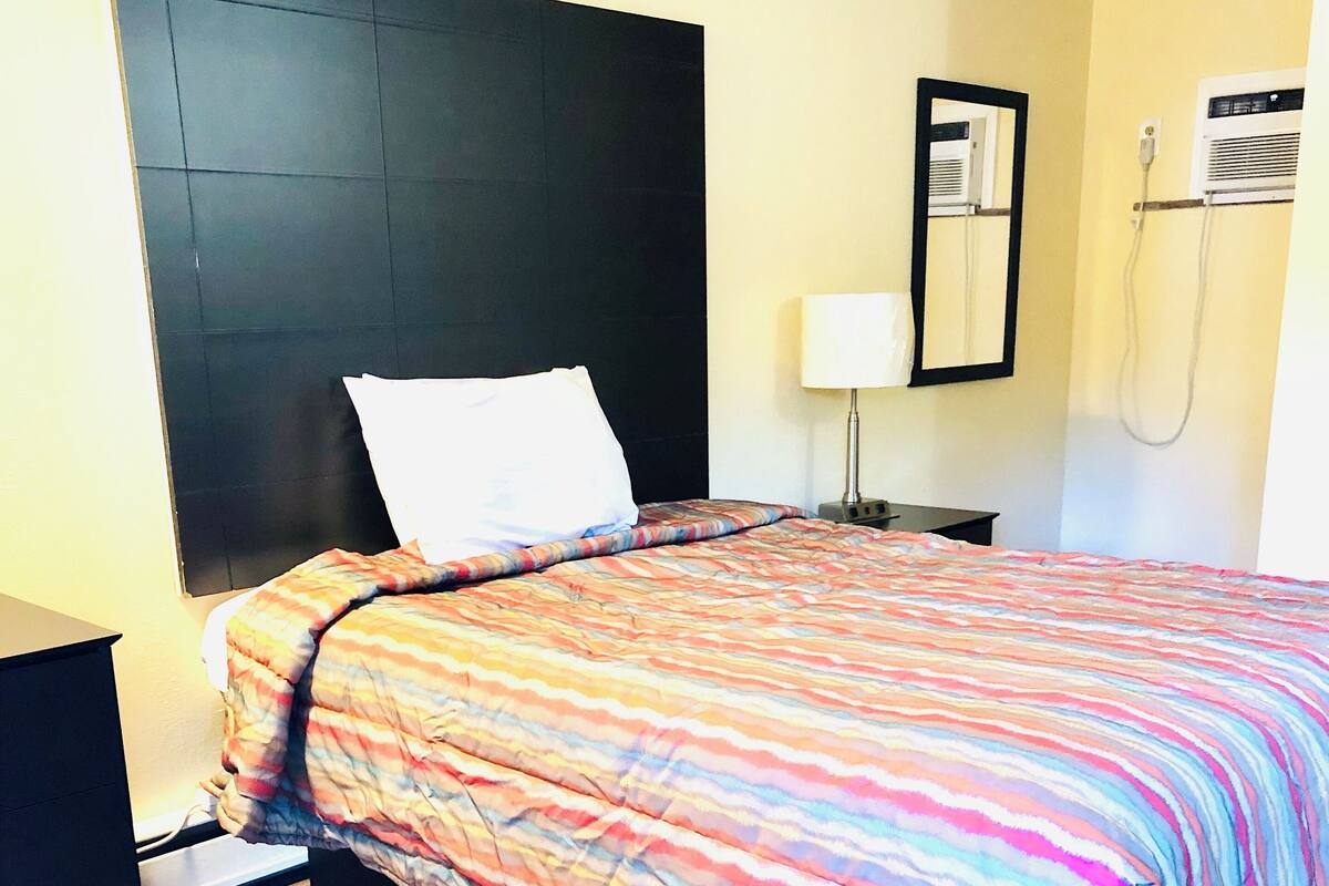 A hotel room with a bed, a nightstand, a lamp, and a wall-mounted air conditioning unit.