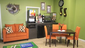 Une chambre d’hôtel avec un mur vert, un canapé, une machine à café, un réfrigérateur, une table à manger avec des chaises et une horloge au mur.