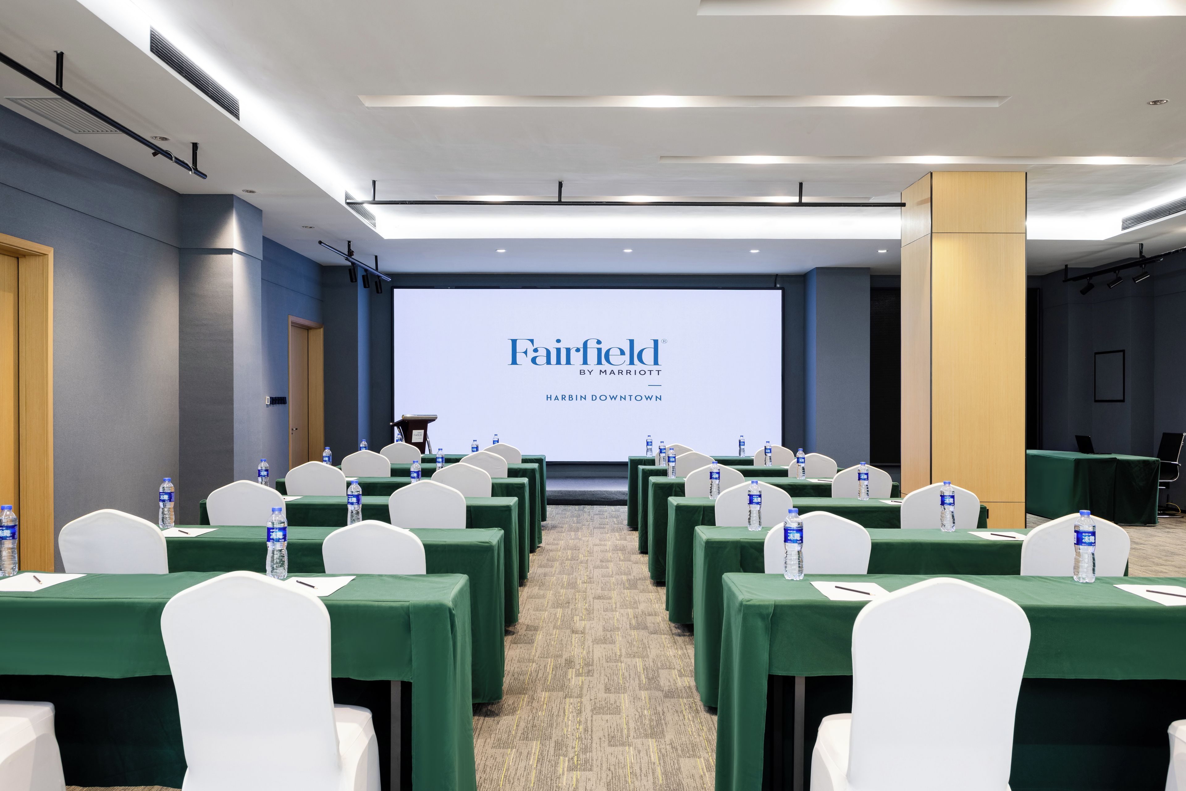 A conference room with rows of tables, chairs, and a presentation screen displaying 'Fairfield by Marriott Harbin Downtown'.