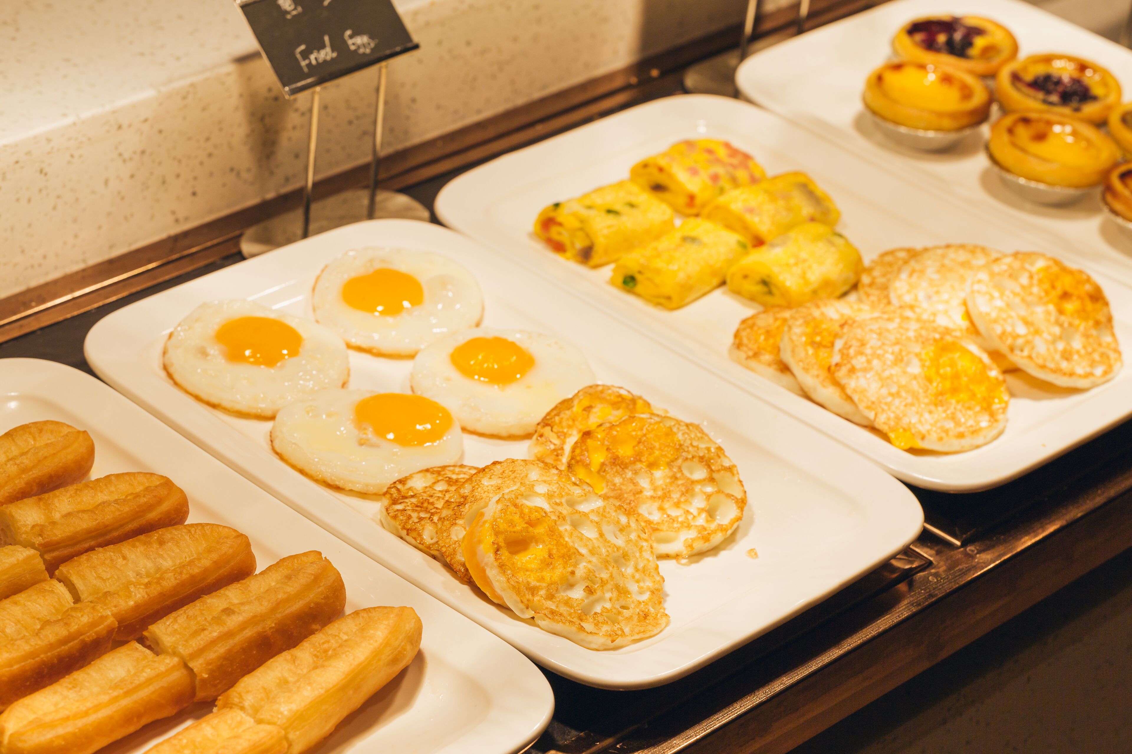 Een ontbijtbuffet met diverse gerechten, waaronder eieren, gebak en custardtaartjes.