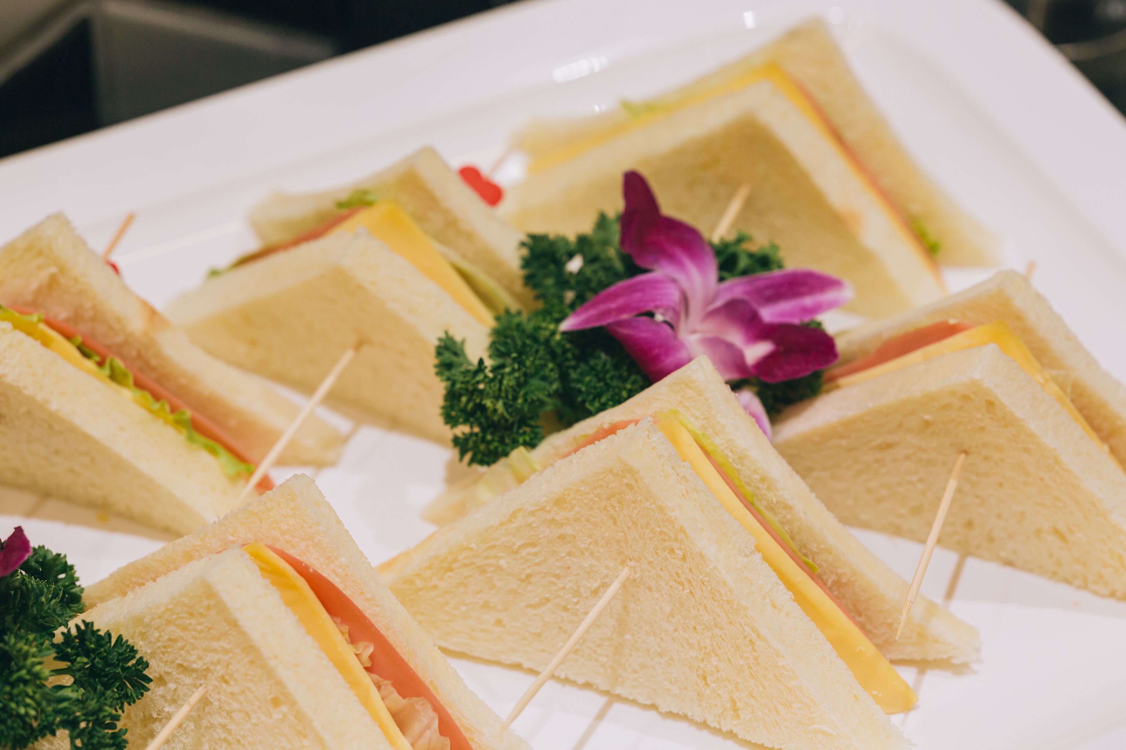 Une assiette de sandwichs triangulaires, maintenus par des cure-dents, et décorés de persil et d’une fleur violette.