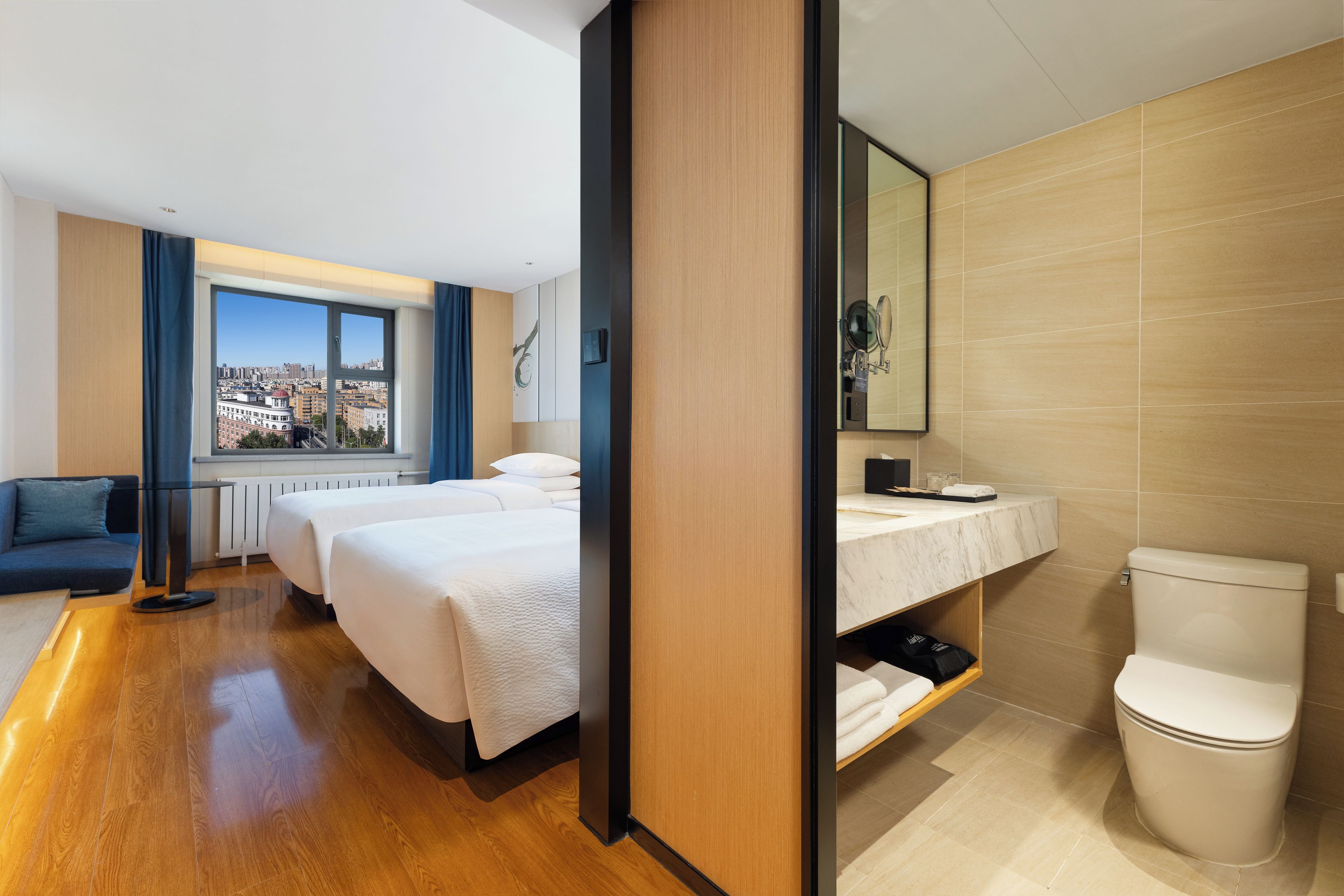 A hotel room with a large window, a bed, a sofa, a bathroom with a toilet, and a mirror.