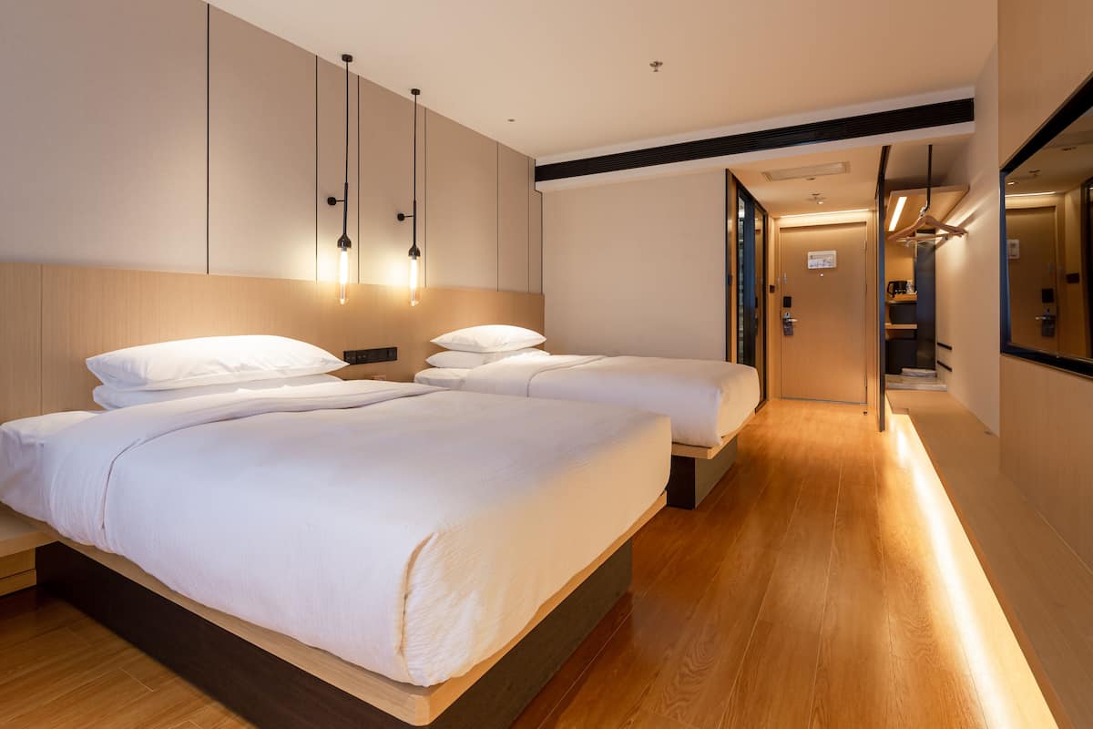 A modern hotel room with two beds, wooden flooring, and a hallway leading to a bathroom.