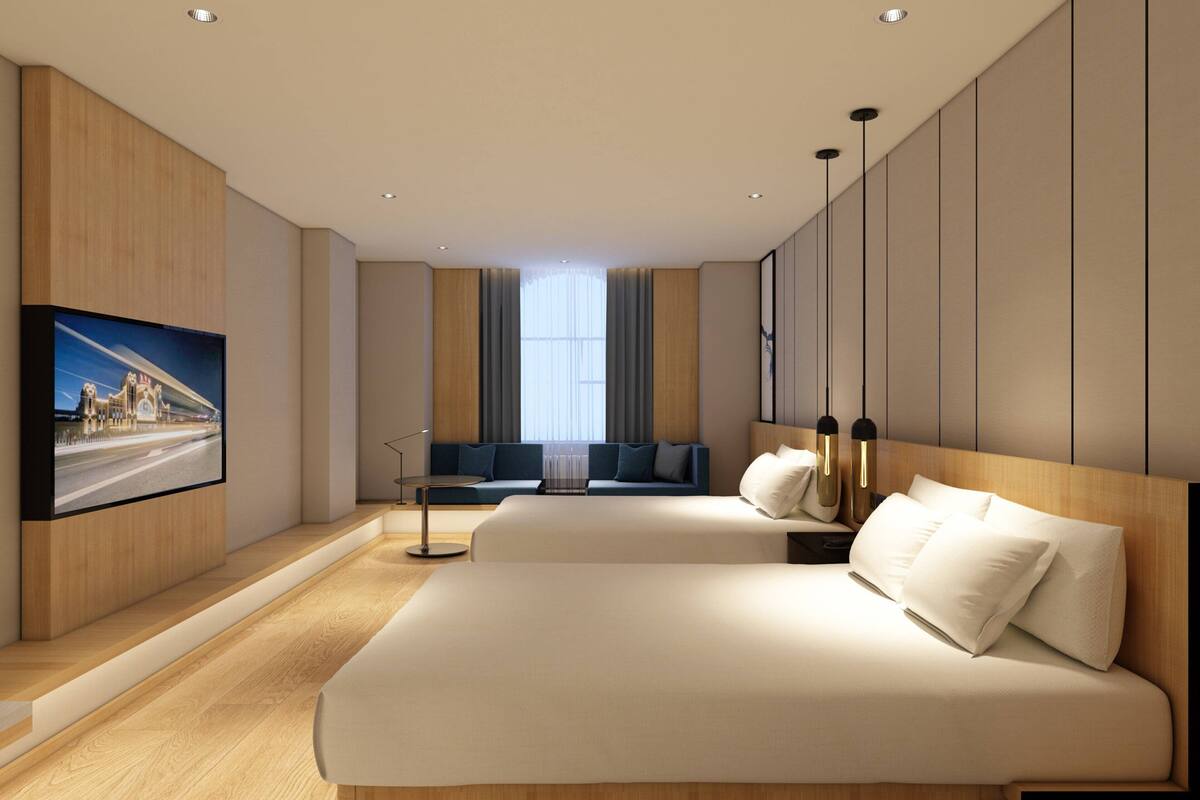 A modern hotel room with a large bed, a seating area with a sofa and chairs, a flat-screen TV mounted on the wall, and a large window providing natural light.
