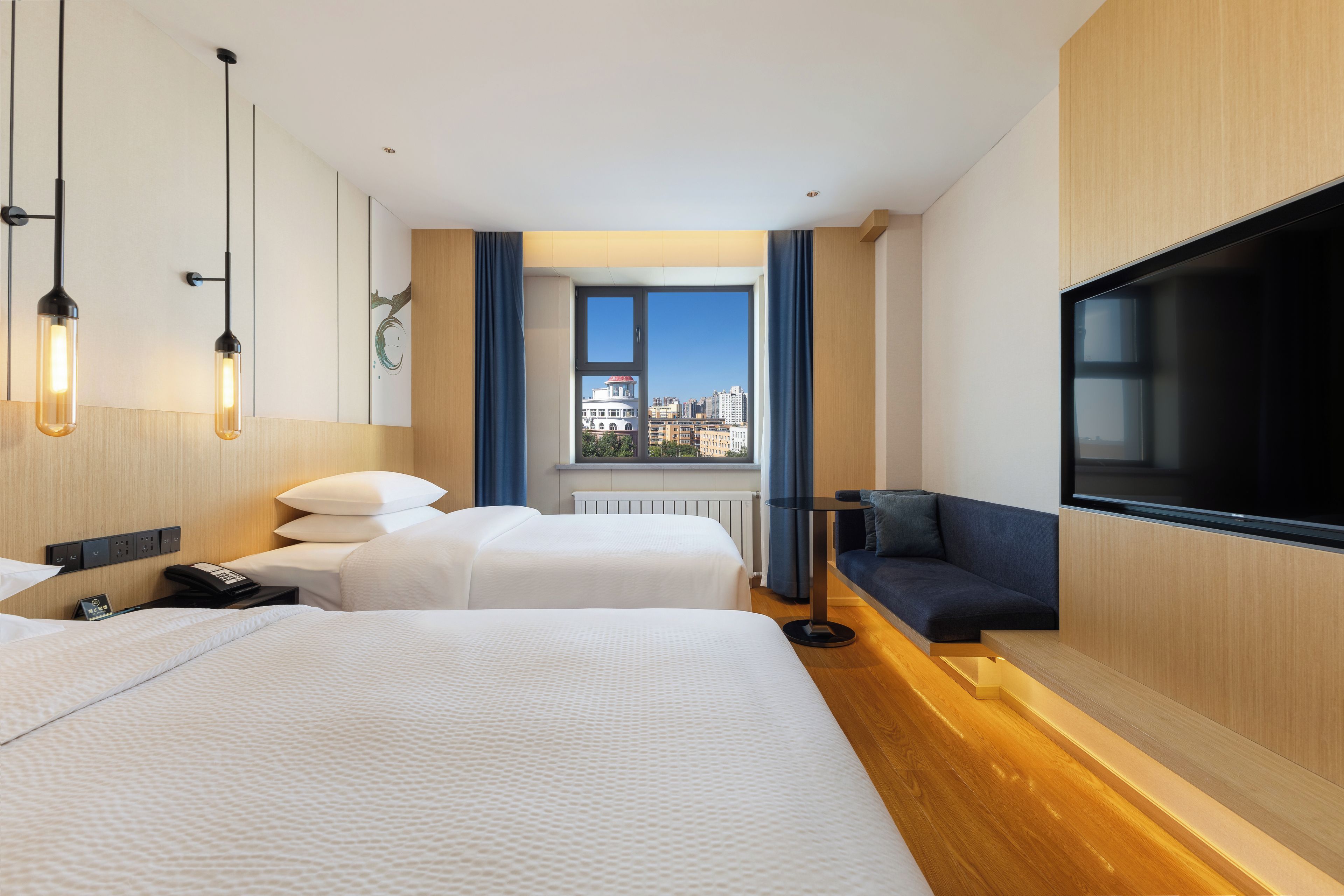 Une chambre d’hôtel avec deux lits, une télévision, une fenêtre donnant sur la ville et un bureau avec une lampe.