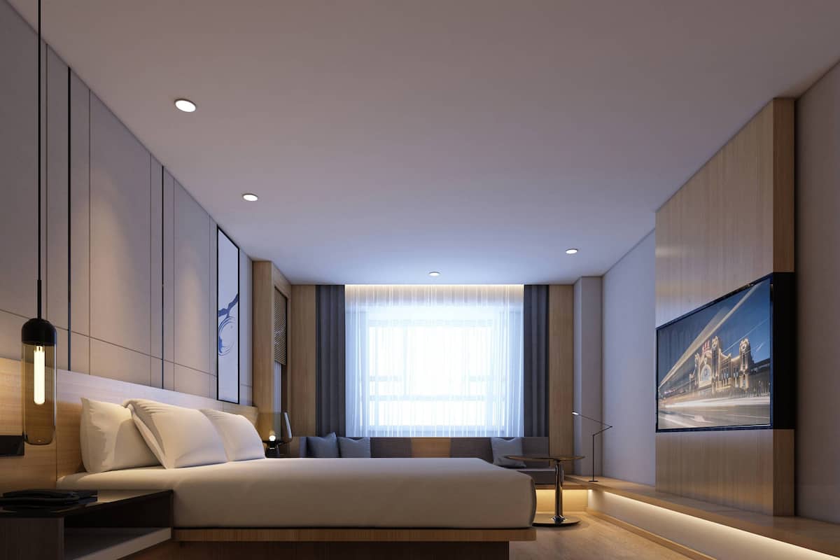 A modern hotel room with a large bed, a flat-screen TV mounted on the wall, and a large window with curtains.