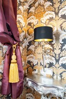 Quarto com papel de parede decorativo, mesinha lateral metálica e abajur com cúpula preta.