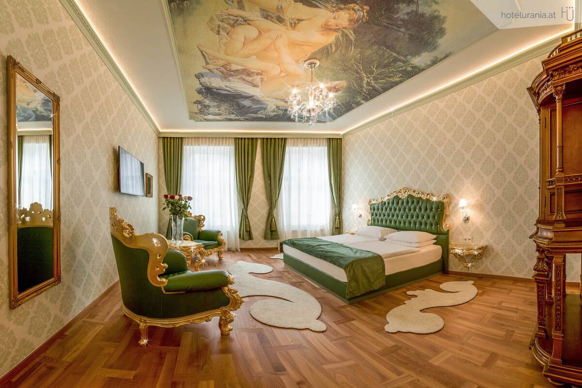 A luxurious bedroom with a large bed, ornate green and gold furniture, a chandelier, and a ceiling mural.