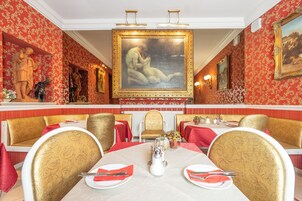 Uma sala de jantar com decoração em vermelho e dourado, incluindo papel de parede estampado, cadeiras douradas e um quadro central.