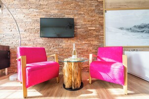 Um quarto com parede de tijolos, televisão montada, mesa de madeira redonda e duas cadeiras rosa vibrante.