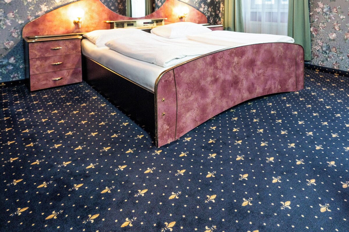 Une chambre à coucher avec un lit à la forme arrondie, un papier peint à motifs floraux, un lustre et une grande fenêtre avec des rideaux.