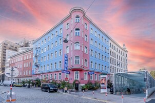Um hotel de vários andares com fachada rosa e azul, situado no encontro de duas ruas e com abrigo de vidro.