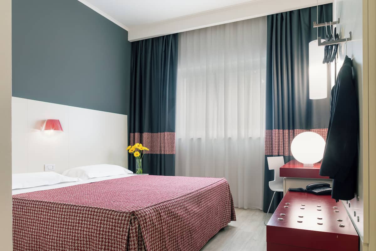A hotel room with a bed, a desk with a mirror, a telephone, and a red storage unit.