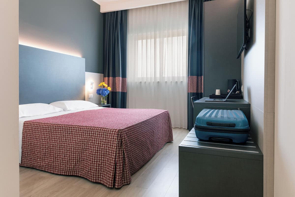 A hotel room with a bed, a desk with a suitcase, a lamp, and a window with curtains.