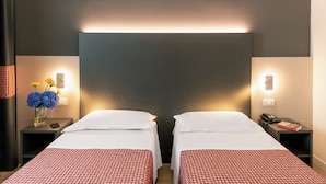 Habitación de hotel con dos camas, una mesita de noche con un teléfono y gafas, un florero con flores y una lámpara.