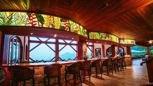 Zona de bar con sillas de mimbre, mostrador de piedra, ventanas amplias con vistas y vigas de madera en el techo.