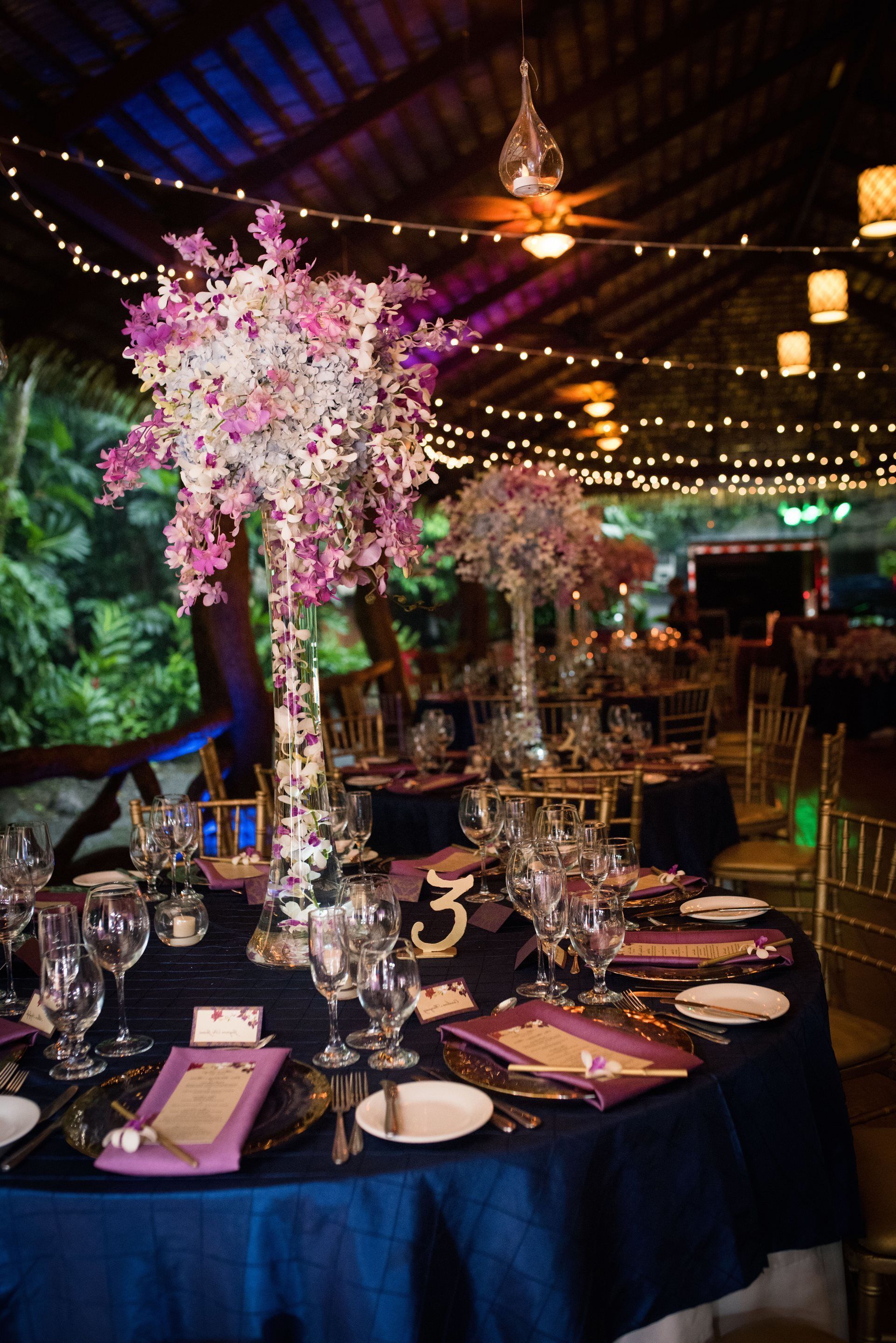 Un salón de banquetes con mesas dispuestas para un evento formal, con centros de mesa florales altos y luces colgantes.