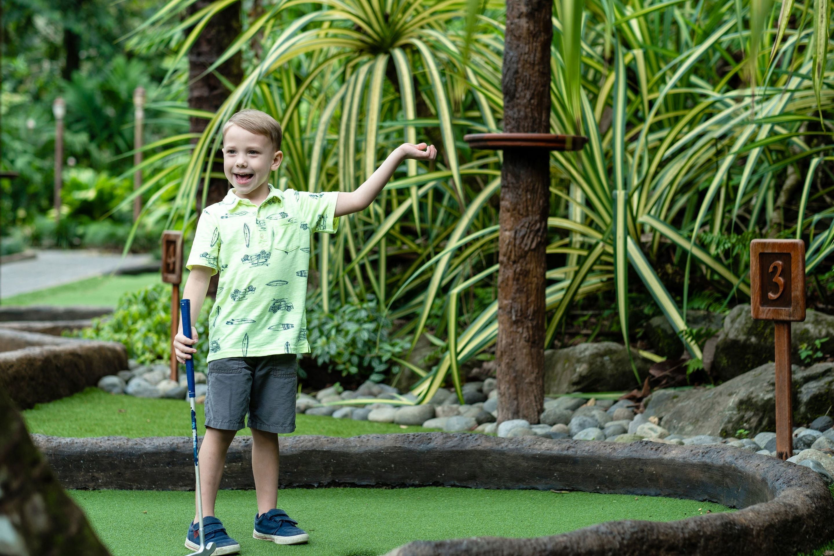 Un niño pequeño sosteniendo un palo de golf en un jardín con un putting green y señales numeradas.