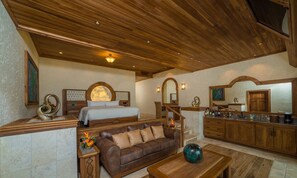 A spacious living area with a wooden ceiling, a large bed, a leather sofa, a coffee table, and a kitchen area with a bar.