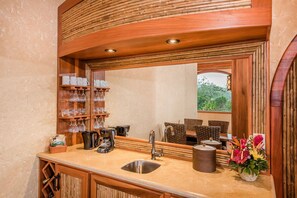 A kitchen with a large mirror, wooden shelves with kitchenware, a sink, and a window with a view of greenery.
