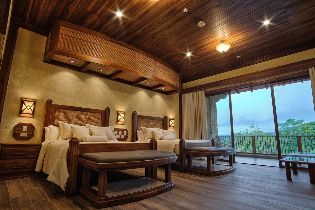 A spacious bedroom with a large bed, a sofa, and a small table. There are two wall-mounted lights and a ceiling light. The room has a wooden ceiling and a large window with a view of trees.