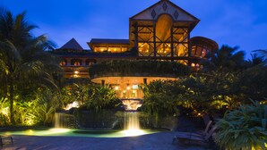 Un hotel grande de varios pisos con una torre central destacada, piscina y exuberante jardinería tropical.