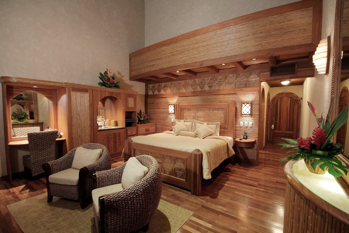 A spacious bedroom with a large bed, wooden headboard, wicker chairs, and a wooden vanity.