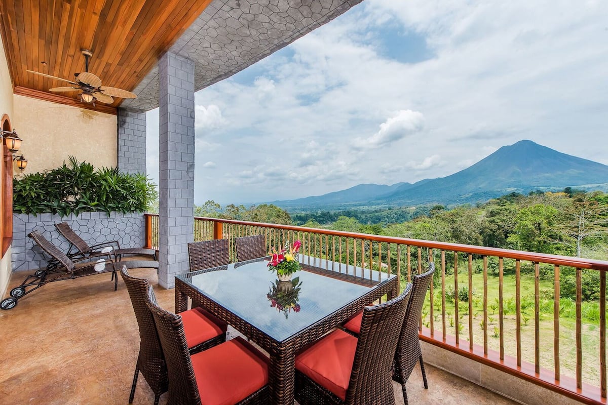 A balcony with wicker furniture, a glass table, and a view of a mountain.