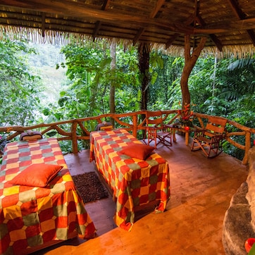 A thatched-roof outdoor seating area with two checkered beds and a stone water feature.