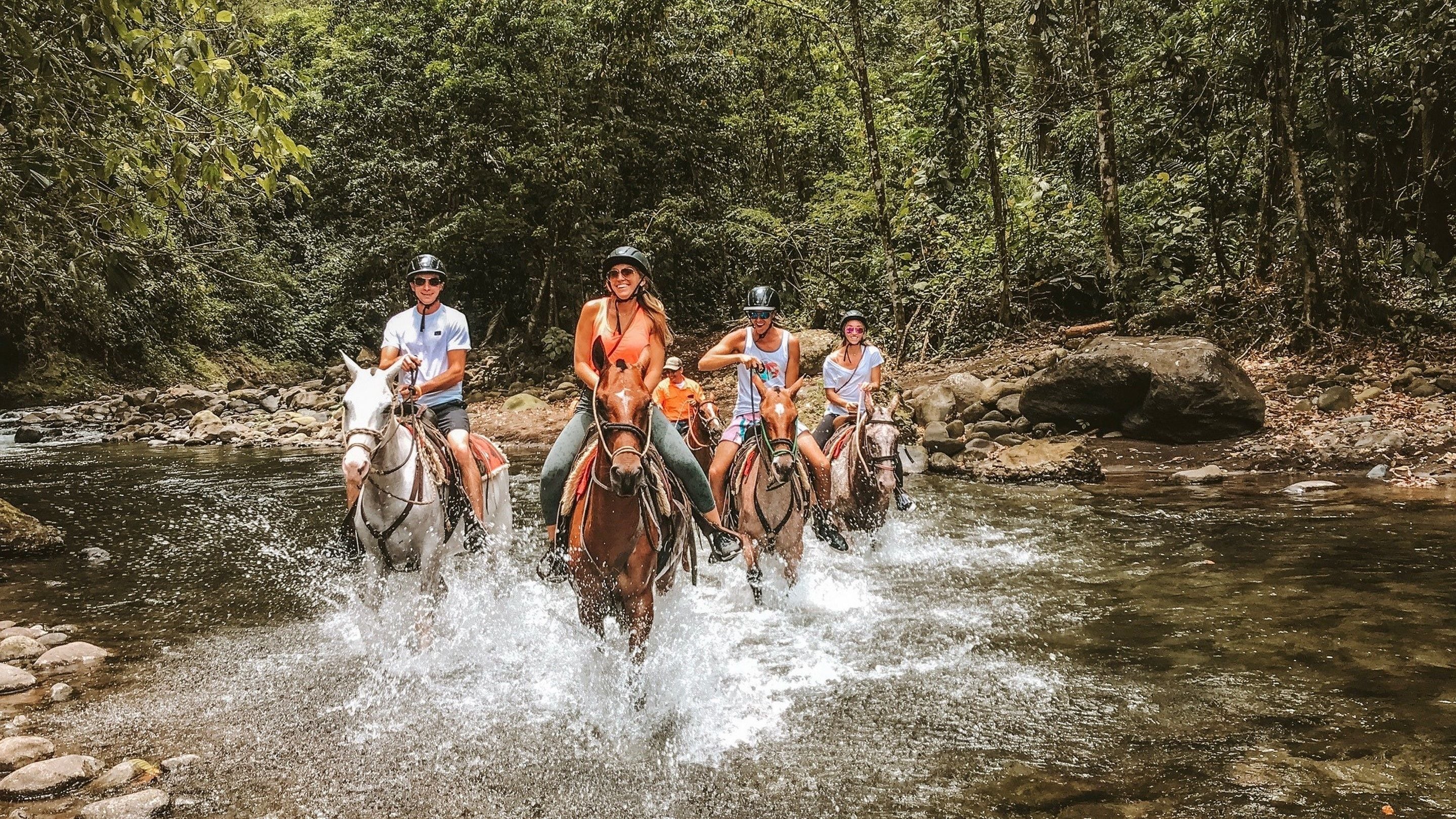 Four people riding horses through a shallow stream surrounded by dense forest.