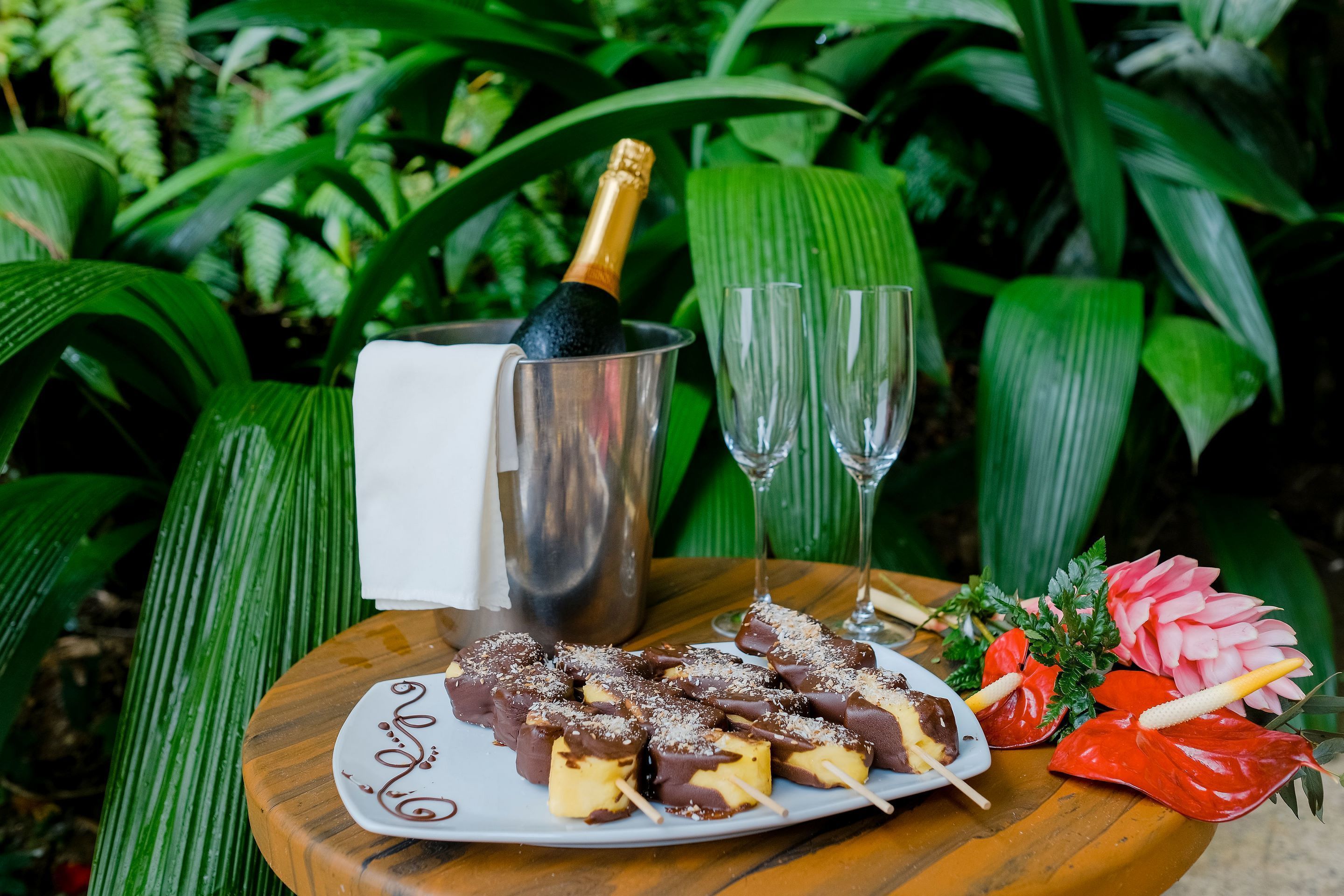 Una mesa de madera con un plato de dulces bañados en chocolate, una botella de champán y dos copas.