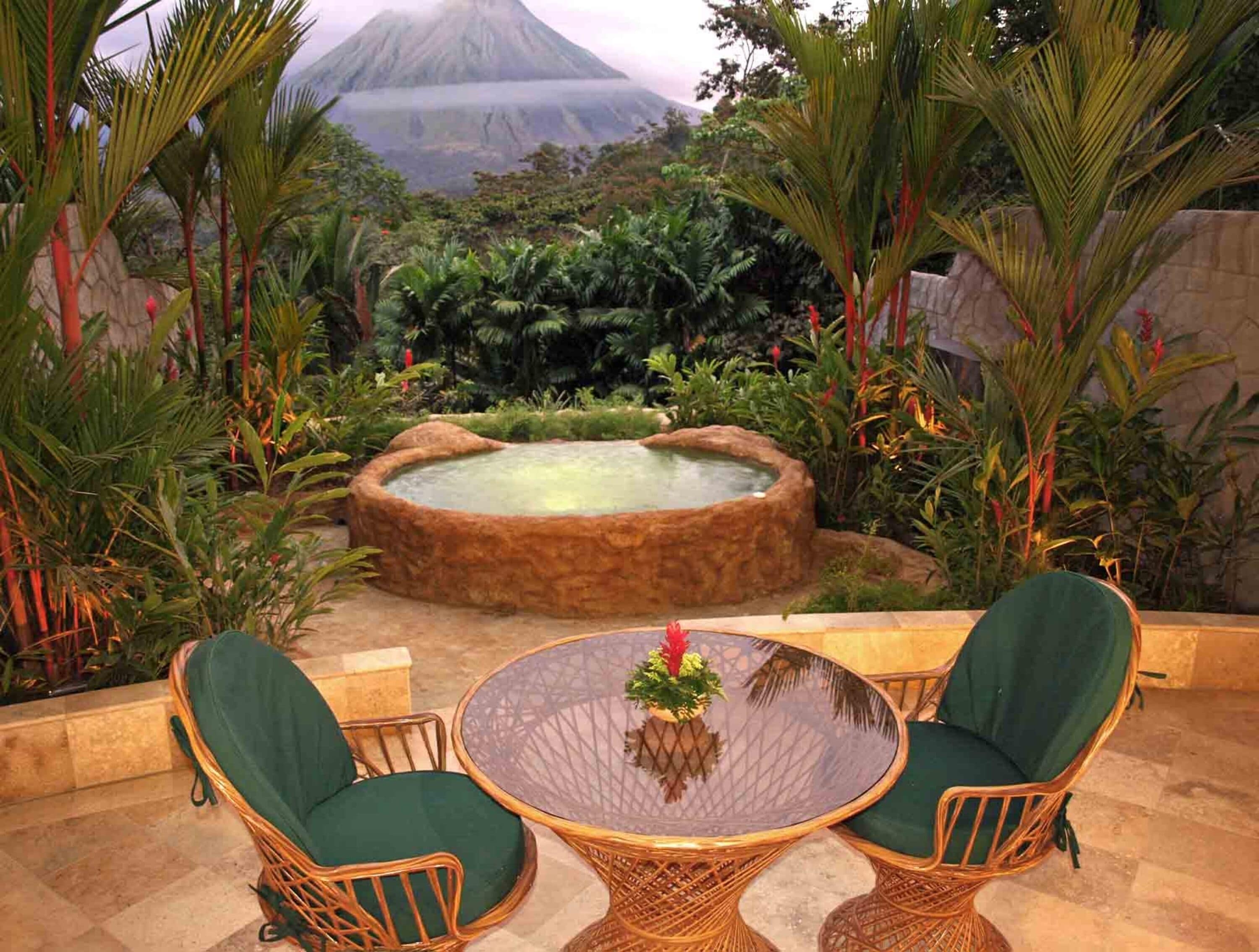 Un espacio al aire libre de estilo tropical con sillas de mimbre y una mesa de vidrio, rodeado de exuberante vegetación y un jacuzzi.