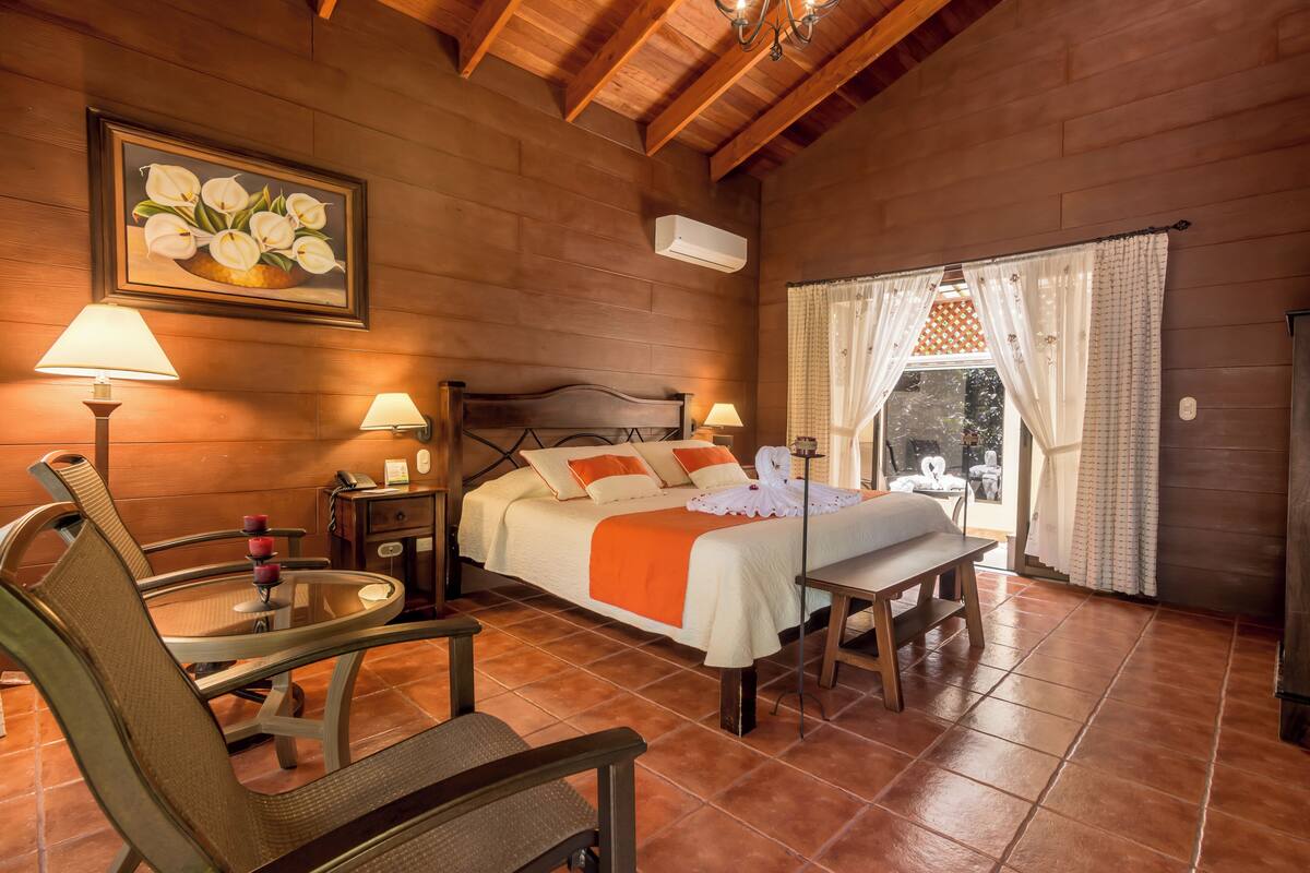 A cozy wooden room with a bed, two bedside lamps, a table with chairs, and a window with curtains.