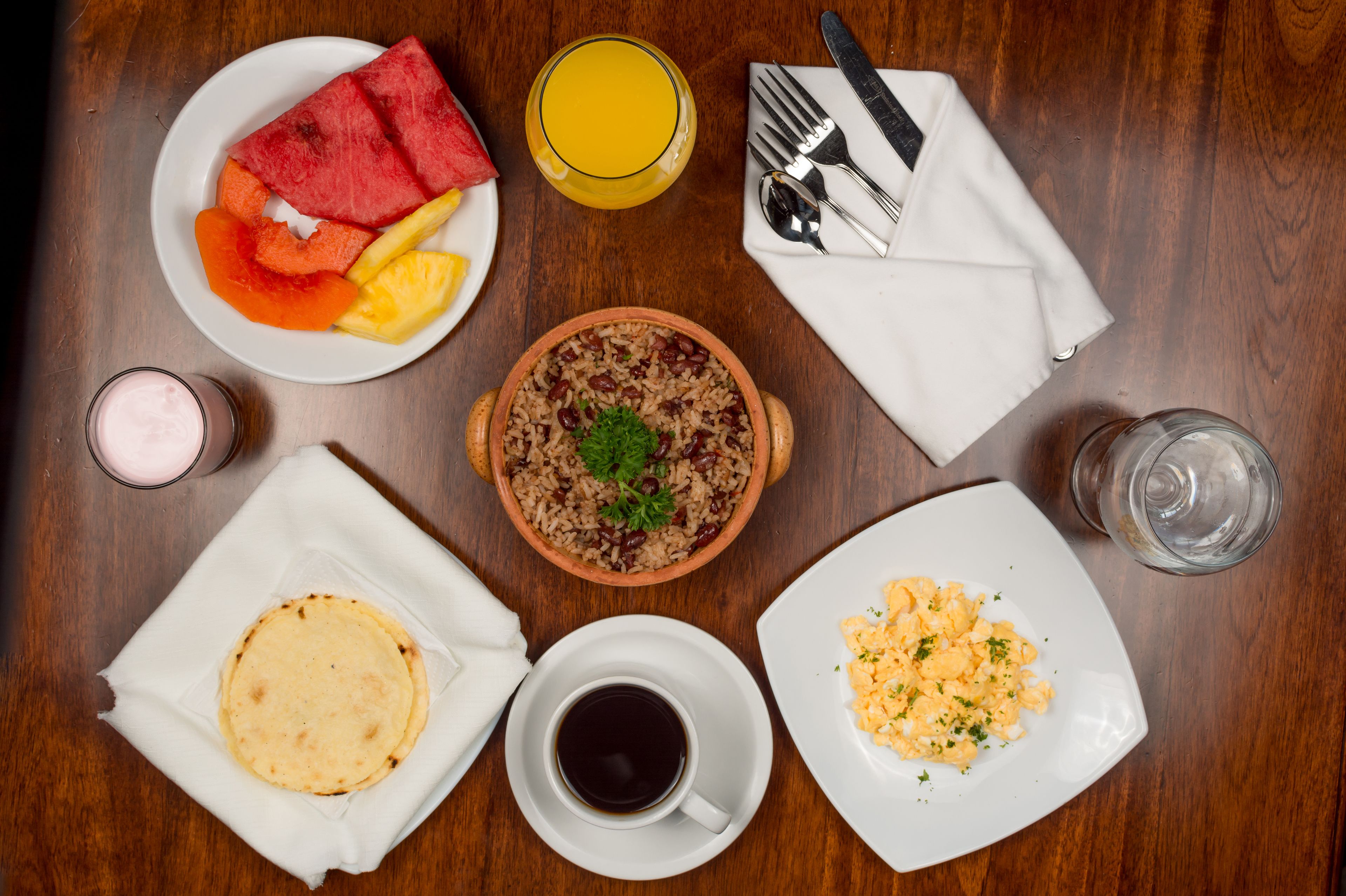 A breakfast setting with a plate of fruit, a bowl of rice, scrambled eggs, a cup of coffee, and a glass of orange juice on a wooden table.