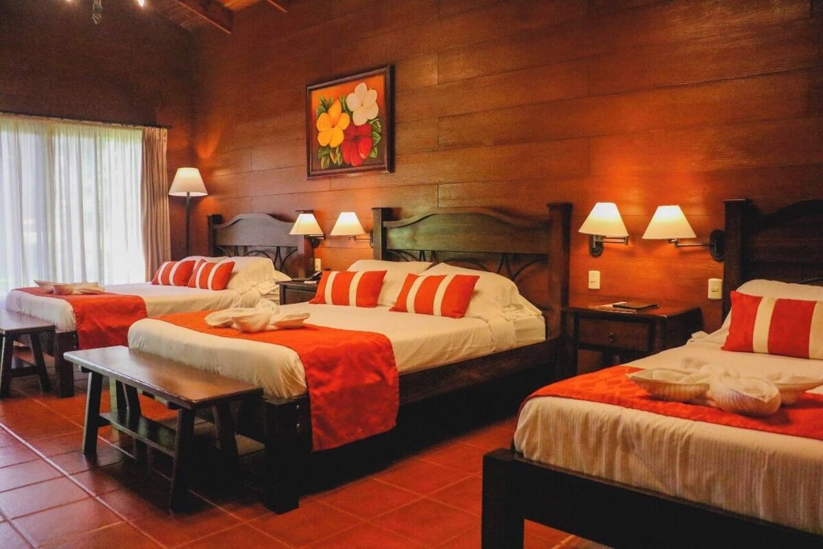 A hotel room with three beds, each with white bedding and orange accents, wooden headboards, and bedside lamps.