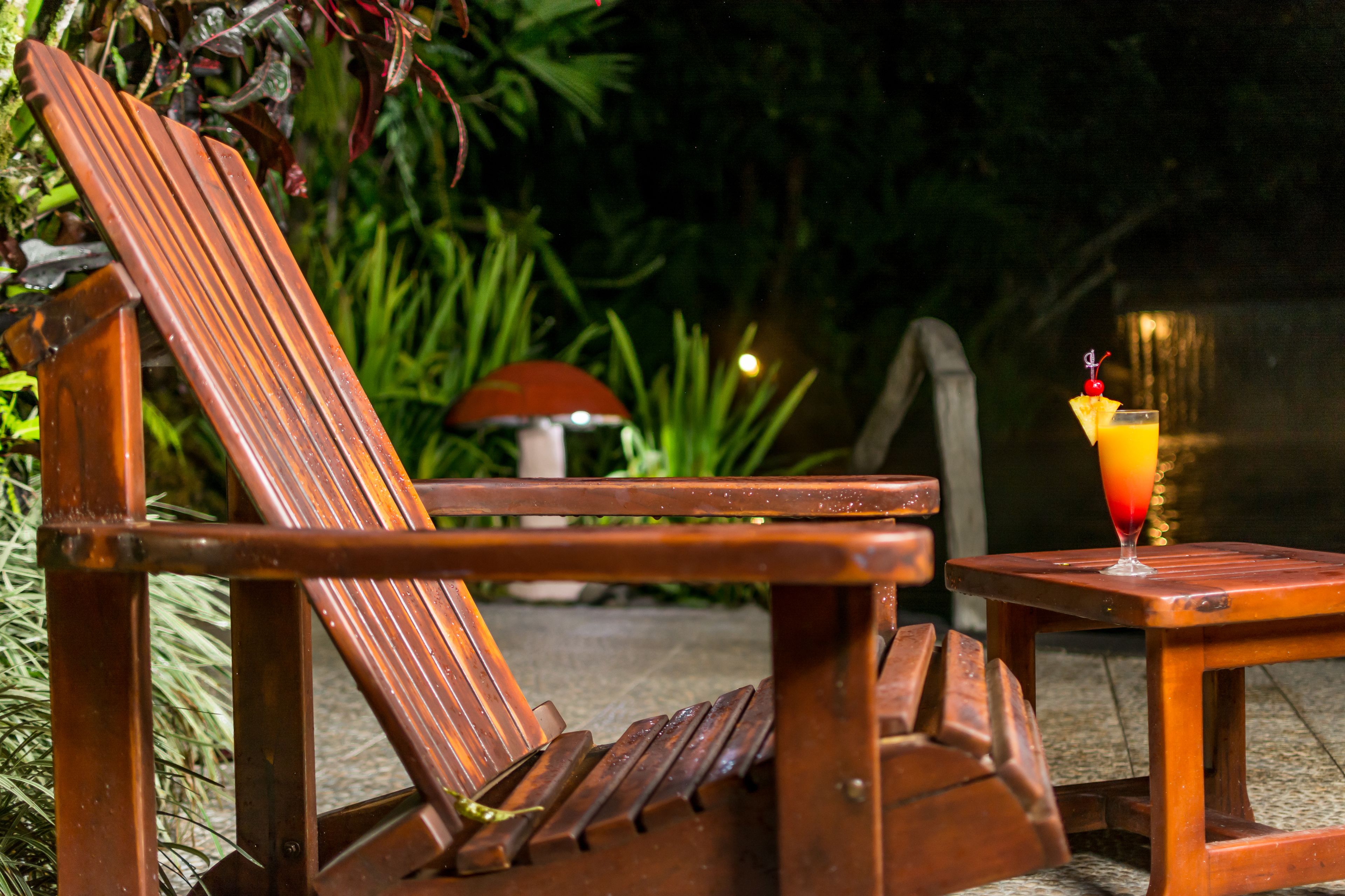 A wooden lounge chair with a small table beside it, a glass of drink with a cherry on the table, and a mushroom-shaped lamp in the background.