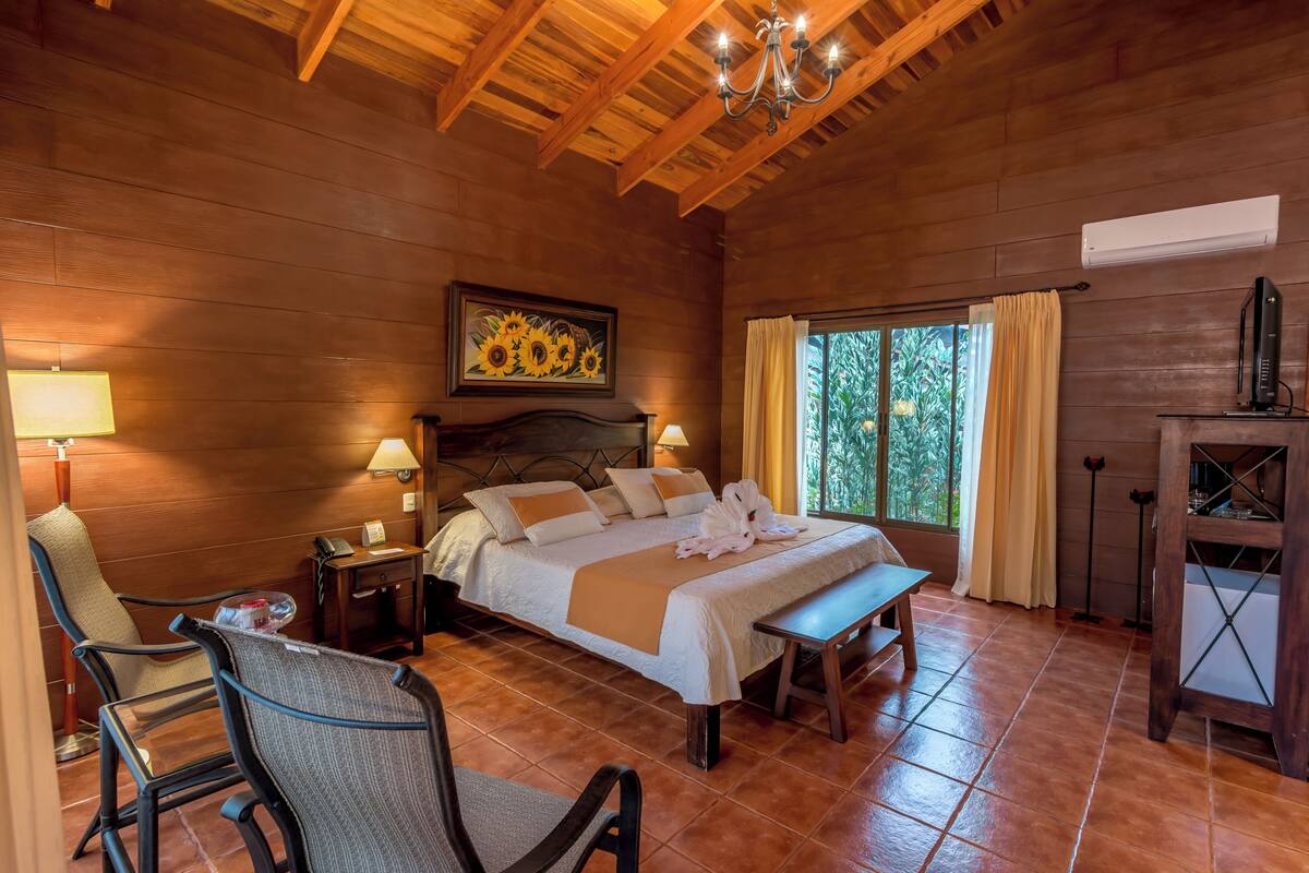A wooden-floored bedroom with a bed, bedside tables, a chair, a bench, and a TV.