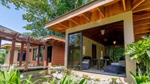 Een modern huis met een glazen deur naar een woonruimte, een houten pergola en een vijver met stenen en planten.