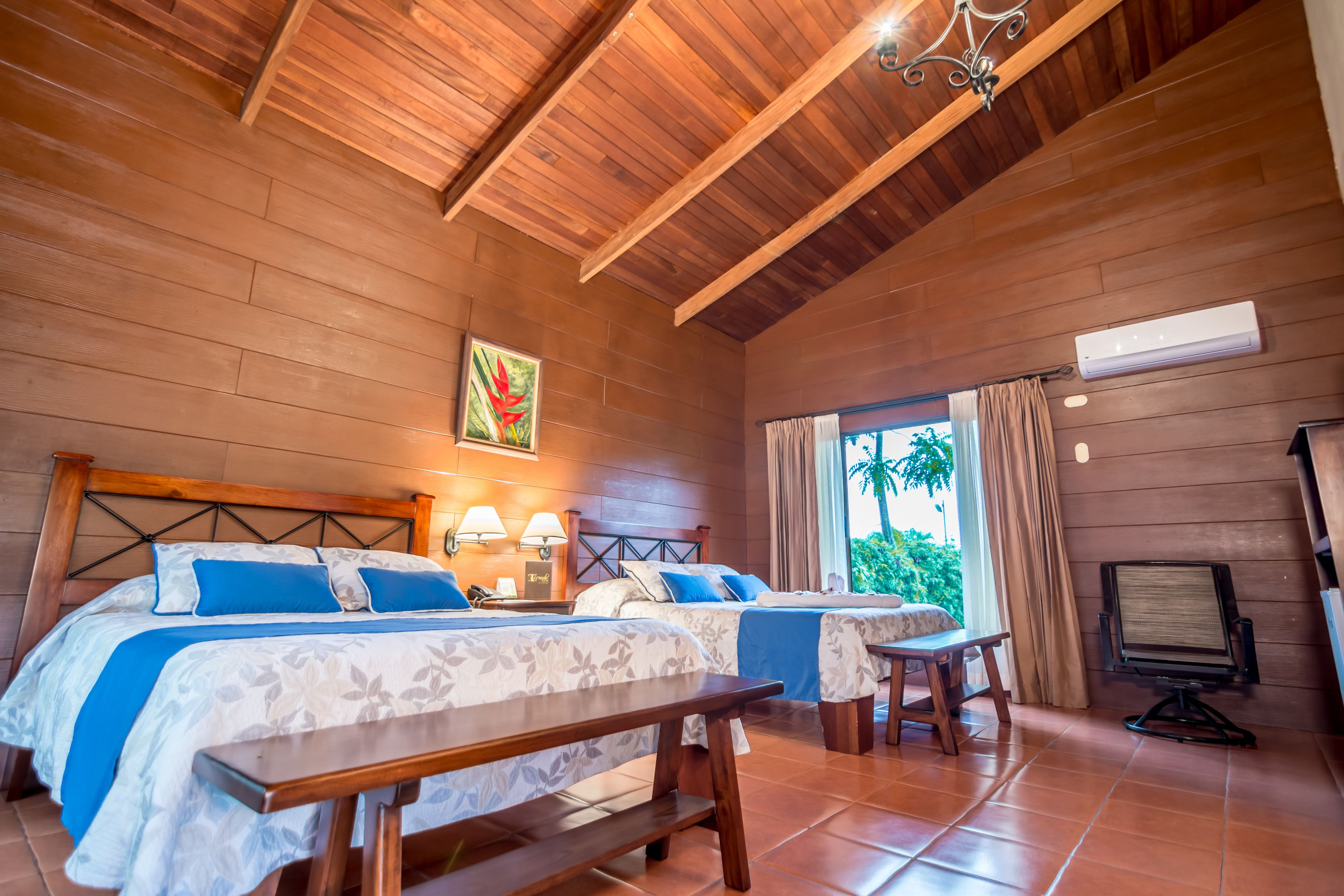 A room with two beds, a wooden ceiling, a window with curtains, and a desk with a chair.