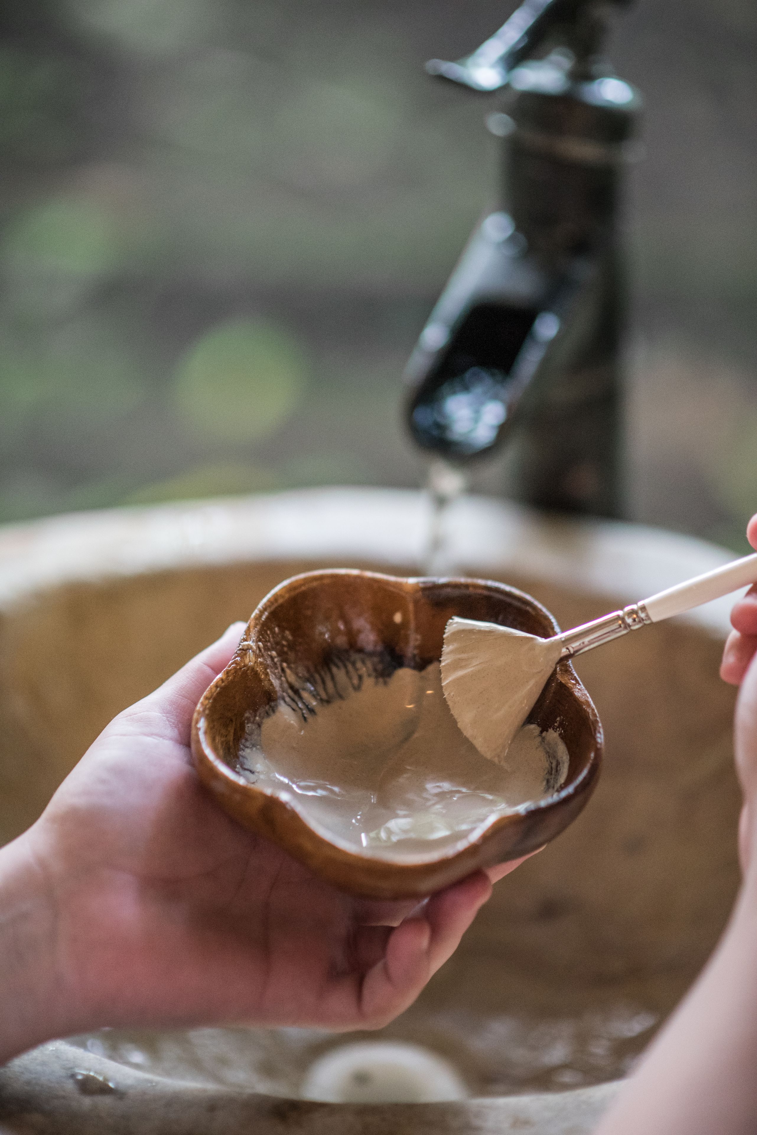 A hand holding a small bowl with a brush, with water flowing from a faucet in the background.