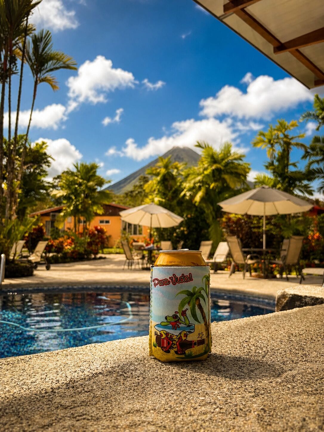 Una lata de cerveza 'Pure Vida!' al lado de una piscina, con vista a una montaña y palmeras.