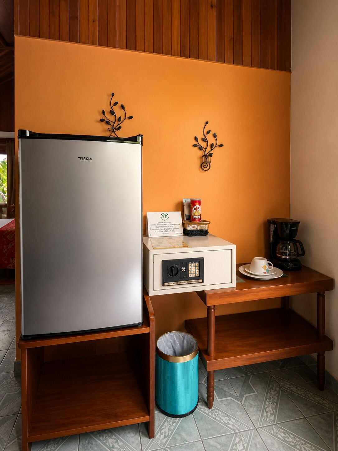 Una kitchenette con refrigerador, cafetera, basurero y una mesa pequeña con una lata de refresco y una caja.