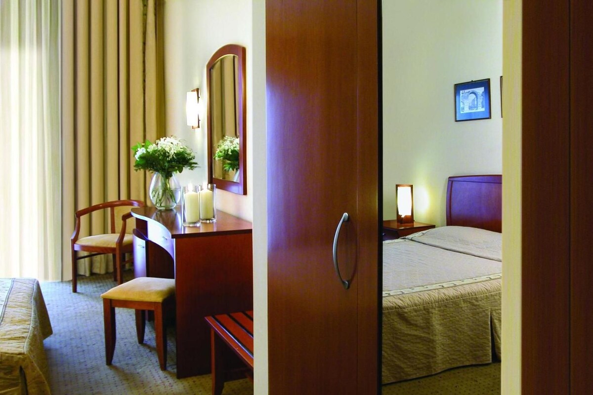 A hotel room with a bed, a desk with a chair, a mirror, and a bedside table with a lamp.