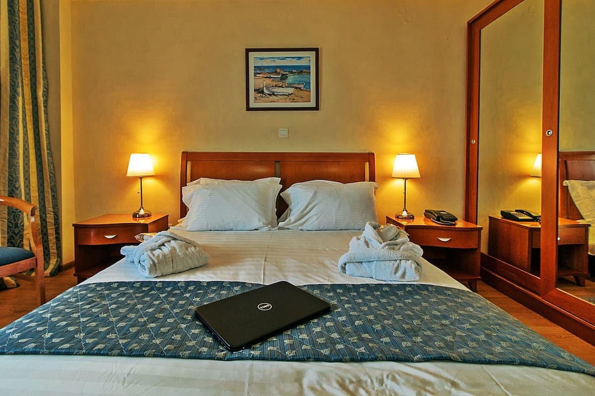 A hotel room with a bed, two bedside lamps, a desk with a telephone, a chair, and a painting on the wall.