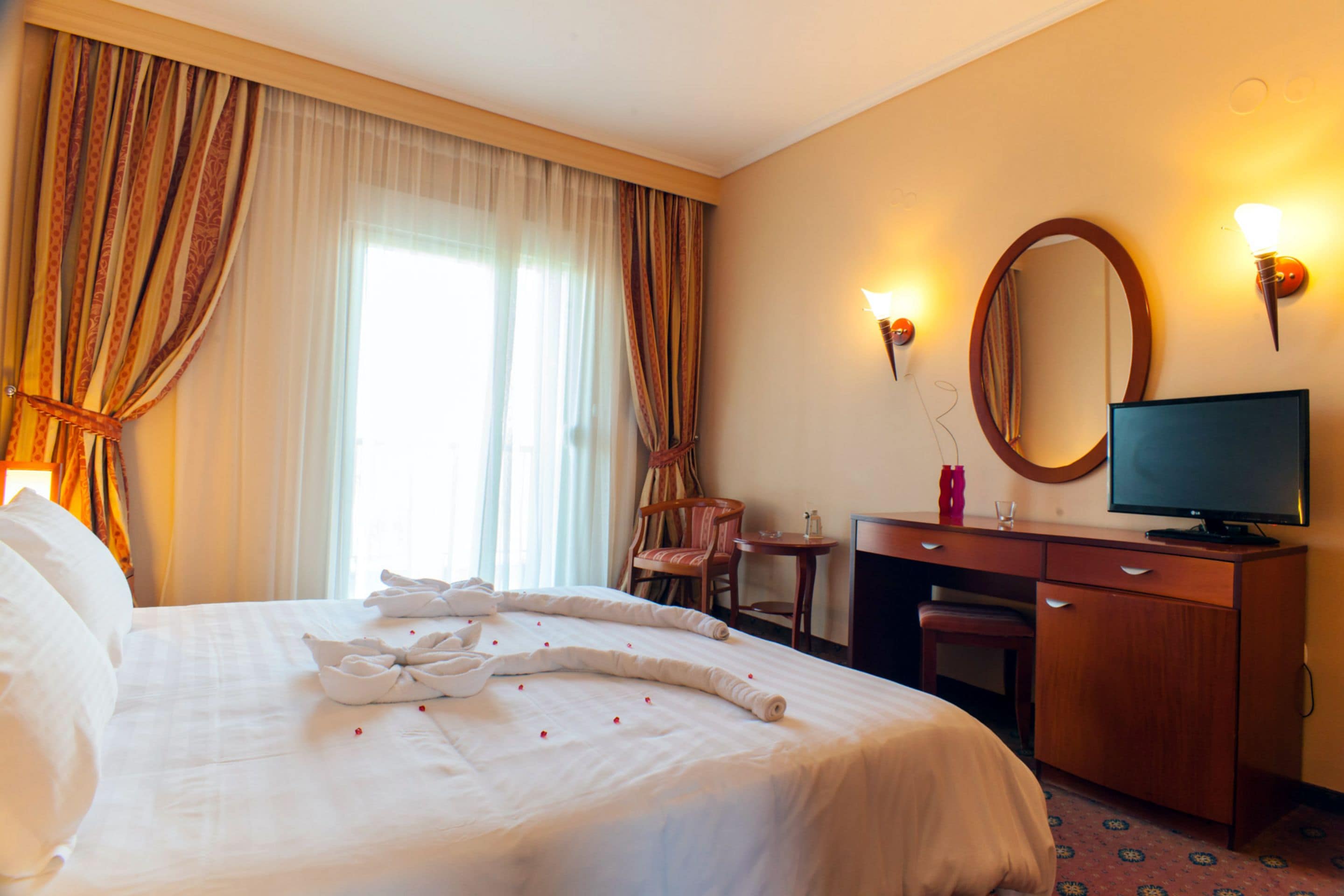 A hotel room with a neatly made bed, a television, a desk with a chair, and a large mirror on the wall.
