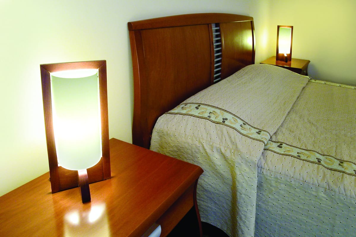 A neatly made bed with a patterned bedspread, a wooden nightstand with a mirror, and a bedside table with a lamp.