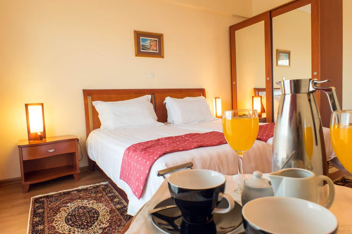 A hotel room with a bed, bedside lamps, a coffee maker, a tray with cups and a pitcher of orange juice, and a patterned rug.
