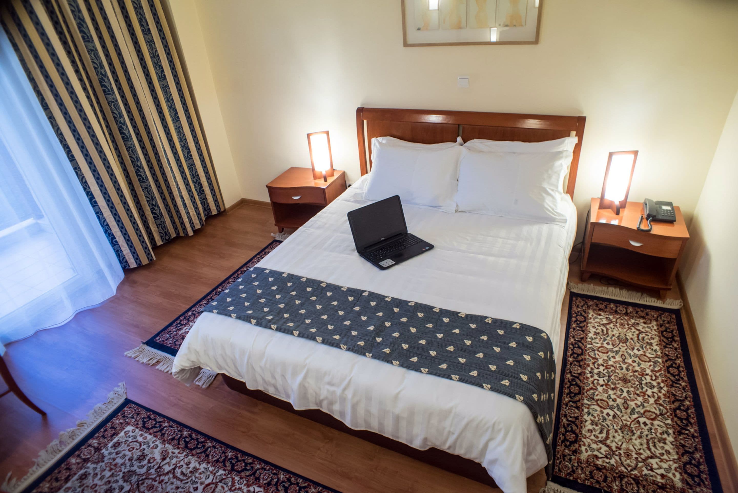 A neatly made bed with a laptop, two bedside tables with lamps, and a patterned rug.