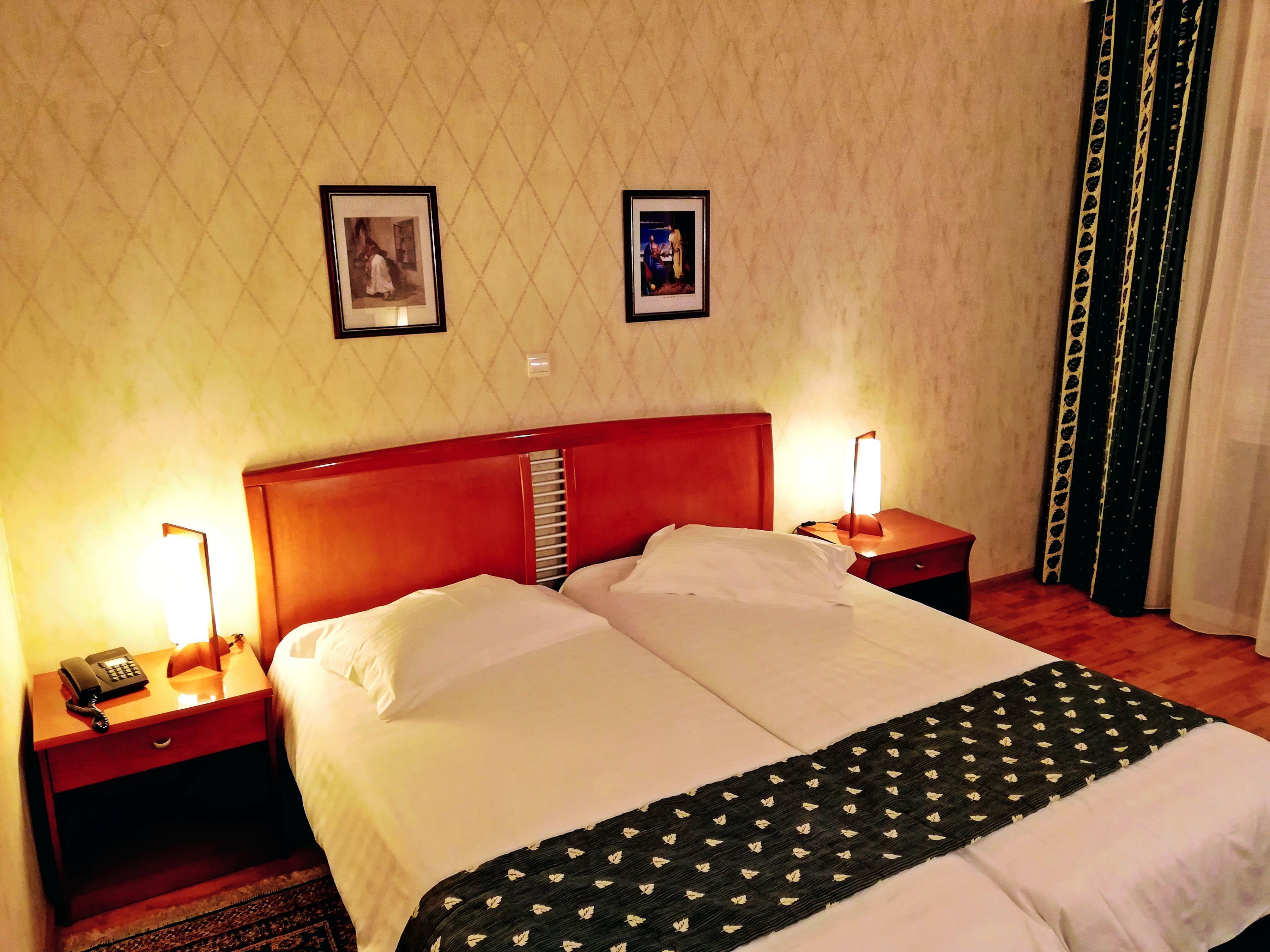 A hotel room with a double bed, two bedside tables, a nightstand, a telephone, and two framed pictures on the wall.