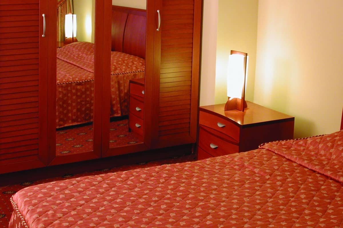 A hotel room with a bed, a nightstand with a lamp, a wardrobe, and a mirror.